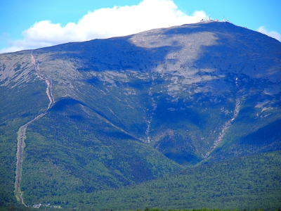 [Close up view of the mountain with an opening in the trees wider than the rail line winding up the left side of the image. Clouds overhead cast shadows on part of the mountain obscuring the view of the rocky top.]